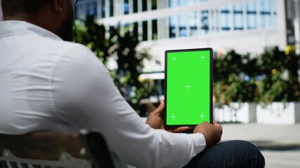 Vertical green screen with black man sitting relaxed on a bench, working online with his tablet near downtown city center. Street style and confident attitude in metropolitan life. Camera B.