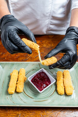 Chef Hands Wearing Black Gloves Breaking Fried Mozzarella Cheese Stick to Show Melted Texture Over Green Plate and Red Sauce