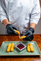 Chef Hands Wearing Black Gloves Breaking Fried Mozzarella Cheese Stick to Show Melted Texture Over Green Plate and Red Sauce
