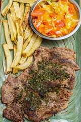 Top View of Grilled Beef Steak Topped with Green Chimichurri Sauce Served with Crispy French Fries and Fresh Tomato Salad