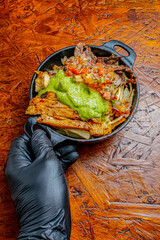 Chef Hand with Black Glove Holding Cast Iron Skillet with Loaded Fries, Grilled Mixed Meat, Guacamole and Pico de Gallo Salsa on Wooden Background