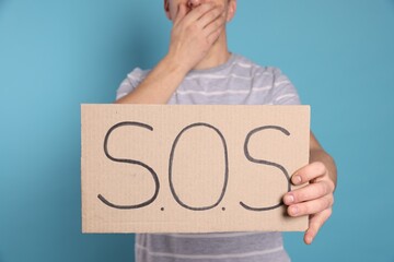 Man holding cardboard sign with word SOS on light blue background, closeup