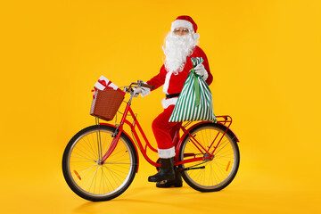 Santa Claus on bicycle with sack and Christmas gifts in basket against yellow background
