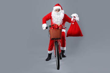 Santa Claus on bicycle with sack and Christmas gifts in basket against gray background