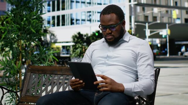 African american man on a park bench scrolling online on his tablet, showcasing relaxed attitude and contemporary street style standing out against the urban scenery. Navigating apps. Camera B.