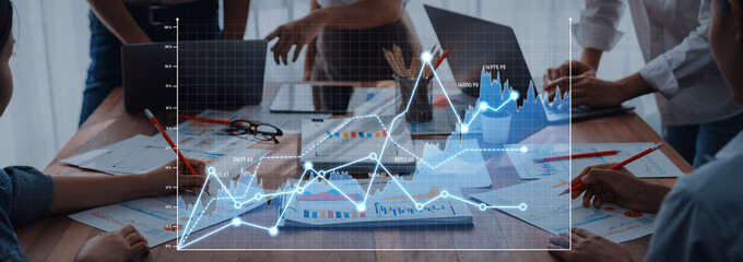 A collaborative business meeting where professionals analyze data trends through graphs on a wooden desk. Laptops and documents create a dynamic workspace for discussions. Scalp
