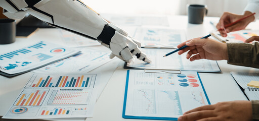A dynamic collaboration scene showcasing human hands and a robot arm analyzing data charts and graphs, illustrating the integration of technology in business strategies.Noogenesis