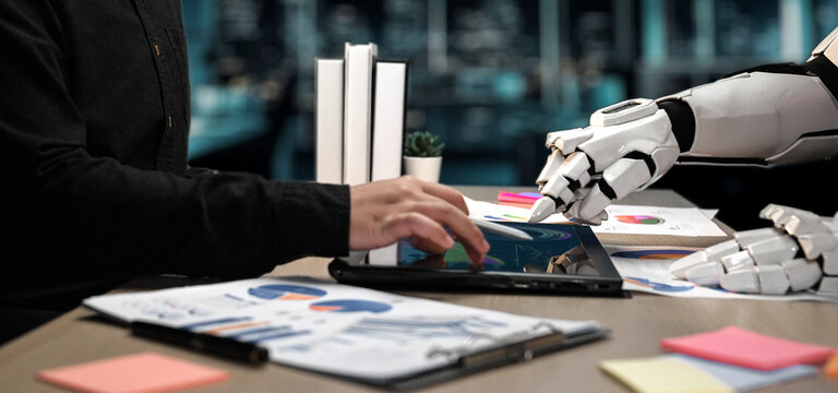 A human hand interacts with a tablet as a robotic arm assists in an office environment. This image illustrates the synergy between human skills and technological efficiency.Noogenesis - Powered by Adobe