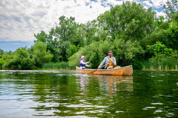 Two people paddle a tandem canoe across calm green water on the Toronto Islands, surrounded by lush summer vegetation, enjoying outdoor recreation and a peaceful escape in nature.