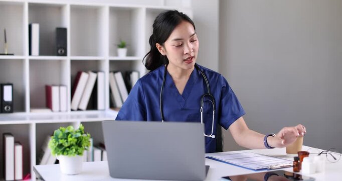 A woman Nurse, in a blue scrubs is smiling while holding a bottle of medicine. She is sitting at a desk with a laptop in front of her. The room is filled with books, some of which are stacked on the d