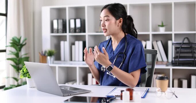 A woman Nurse, in a blue scrubs is smiling while holding a bottle of medicine. She is sitting at a desk with a laptop in front of her. The room is filled with books, some of which are stacked on the d