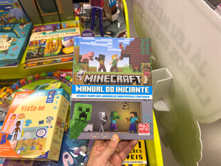 Fototapeta premium BELO HORIZONTE, MG, BRAZIL, DECEMBER 12, 2025 A person's hand holds a vibrant Minecraft Beginner's Manual book, showcasing popular gaming culture in a bright bookstore aisle.