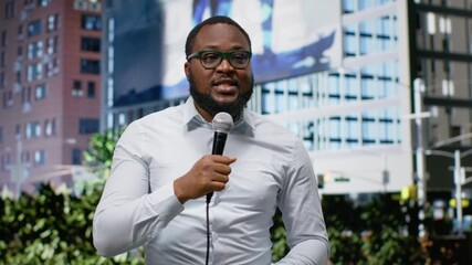 Black presenter in a modern city center speaking into a microphone, using communication and reporting skills to deliver news statement and cover public event in a metropolitan setting. Camera B.