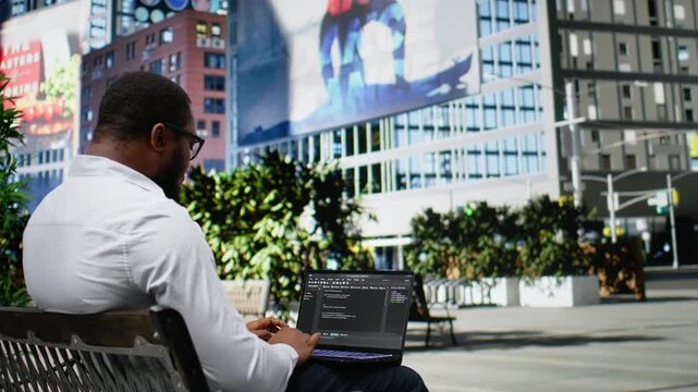 African american guy works on programming code on terminal window, sitting on a bench downtown and working on IT with his laptop in sunlight. Professional attitude in a dynamic city life. Camera A.