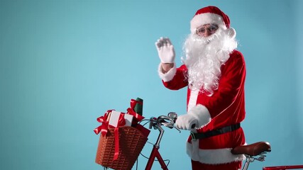 Santa Claus on bicycle with Christmas gifts in basket greeting someone and walking away against light blue background