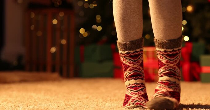 Woman in cozy socks against Christmas tree at home, closeup