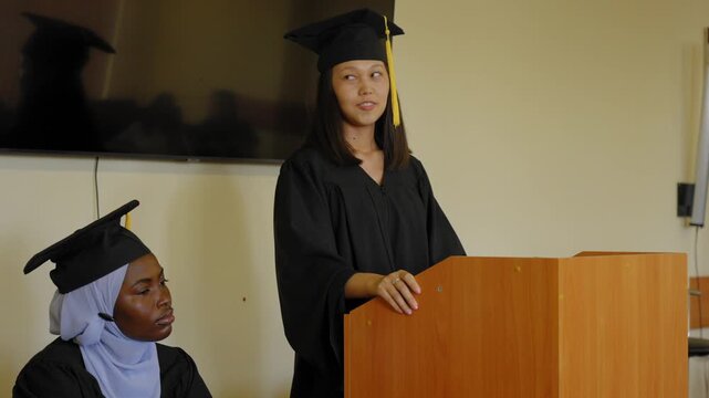 A young woman stands at the lectern and delivers a graduation speech to her classmates. 