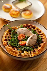 A delicious savory waffle sits on a rustic wooden table, topped with fresh arugula, juicy tomatoes, and a rich creamy filling, inviting the senses in a warm cafe atmosphere