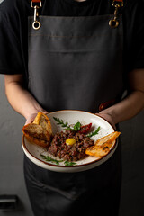 A chef holds a beautifully plated dish featuring savory meat, topped with an egg yolk, paired with golden toast. The background reflects a warm, inviting atmosphere