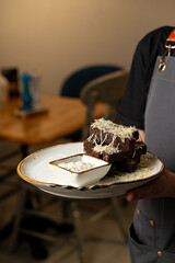 A plate piled high with rich chocolate cake slices sits under a delicate layer of creamy topping. The warm ambiance of a cafe invites all to indulge in this sweet treat