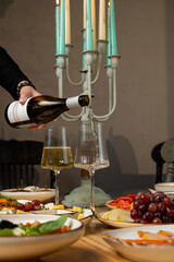 Hands gently pour white wine into crystal glasses while a beautifully arranged dinner table showcases a feast of fresh fruits, cheeses, and delectable snacks, illuminated by soft candlelight