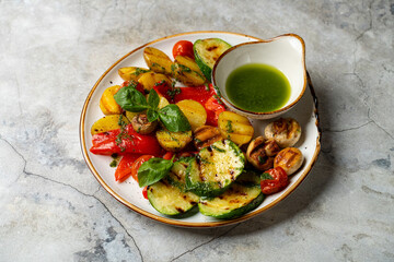 Colorful grilled vegetables including zucchini, peppers, and potatoes are arranged beautifully on a plate, complemented by fresh basil and a bright green dipping sauce