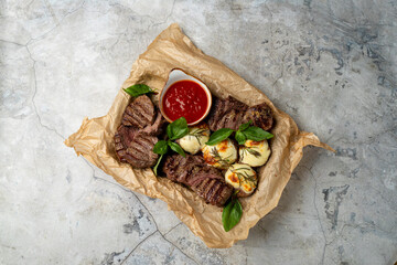 Grilled steak slices are arranged on natural paper, accompanied by creamy stuffed vegetables and a bright red dipping sauce. Fresh basil adds a touch of color and flavor to this hearty meal