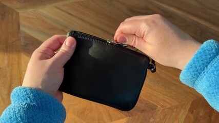 Close up video of hands placing a diamond engagement ring into a black leather wristlet pouch and zipping it closed in natural sunlight on a wooden surface.