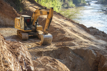 Excavator digging soil steep riverbank next to flowing water, powerful heavy machinery working dangerous terrain, massive yellow construction equipment moving earth near river, industrial scene © CoreRock