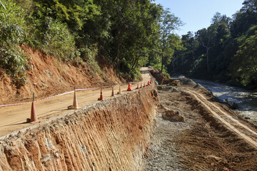View of dangerous dirt road under construction on hillside near river with orange traffic cone markers for safety during daytime in rural forest environment showing development progress