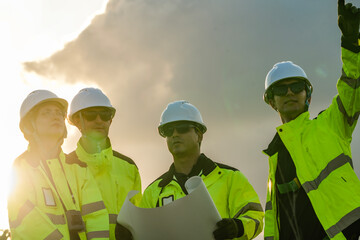 Team of four engineers in white hard hats and high-visibility gear standing at a wind farm at sunset. Professional crew planning low-carbon green energy projects.