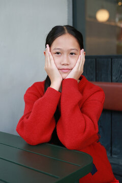 Portrait sweet young Asian girl in red sweater resting her face on her hands while sitting at table indoors.