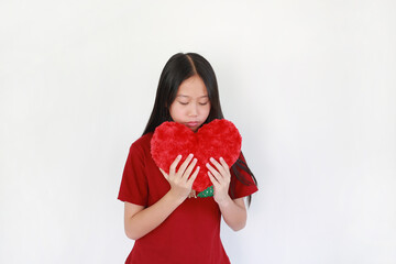 Portrait young Asian girl in red shirt gently holds a large, fluffy red heart pillow to her chest with a sweet, closed-eye expression against white background.