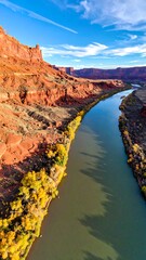 A river cuts through reddish mesas under a bright blue sky with scattered clouds, framed by autumn foliage