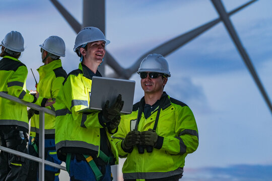 Team of engineers in hard hats and high-visibility jackets using a laptop at a wind farm. Professional technicians collaborating on green energy maintenance at dusk. - Powered by Adobe