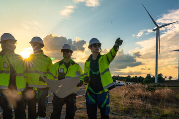 Team of four engineers in white hard hats and high-visibility gear pointing at wind turbines during sunset. Professional crew collaborating on a green energy project for low-carbon power.