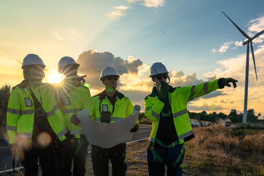 Team of four engineers in white hard hats and high-visibility gear pointing at wind turbines during sunset. Professional crew collaborating on a green energy project for low-carbon power. - Powered by Adobe
