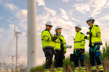 Team of four engineers in white hard hats and high-visibility gear pointing at wind turbines during sunset. Professional crew collaborating on a green energy project for low-carbon power.
