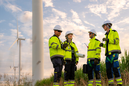 Team of four engineers in white hard hats and high-visibility gear pointing at wind turbines during sunset. Professional crew collaborating on a green energy project for low-carbon power.