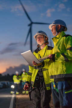 Male and female engineers in safety gear inspecting wind turbines at dusk. Female engineer holding a walkie-talkie while her colleague uses a laptop for green energy monitoring.