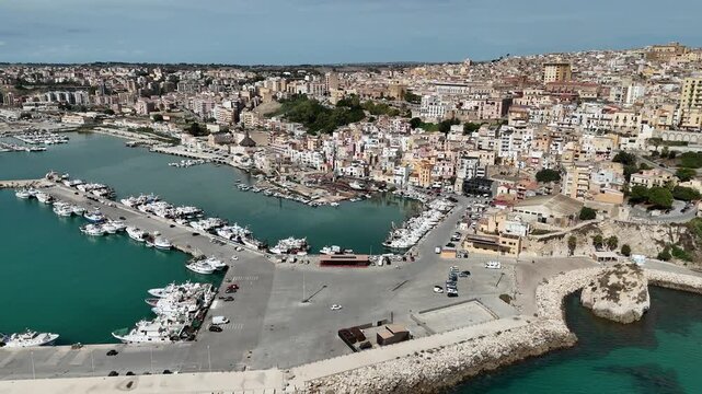 Aerial pullback over Sciacca harbor, descending to reveal the marina, fishing boats, curved breakwater, and dense hillside town along Sicily&rsquo;s southern coast.