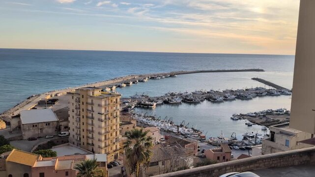 Sunset view over the Sciacca marina, with fishing boats, calm harbor water, warm evening light, and coastal buildings overlooking the port in western Sicily.