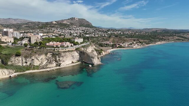 Rotating aerial footage approaching the Sicilian shoreline, revealing turquoise water, limestone cliffs, and coastal towns stretching along the Mediterranean.