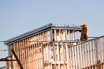 A construction worker installing metal framing and roof supports on a commercial building, illustrating elevated structural assembly, exterior construction progress, and job site safety practices