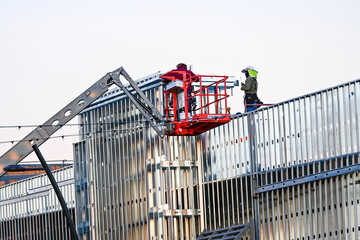 Two construction workers installing metal framing from a boom lift on a commercial building exterior, illustrating coordinated elevated access work, structural assembly, and jobsite safety practices