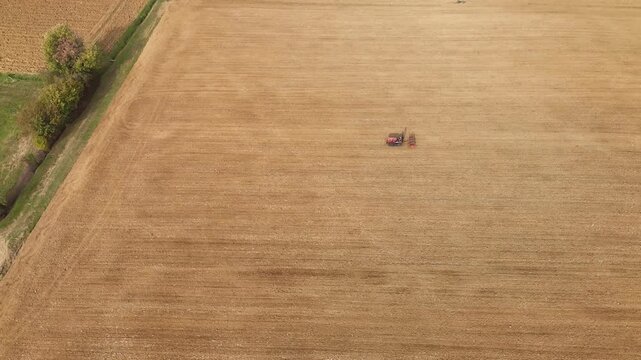 High-angle drone shot of a small tractor working in the middle of a vast golden plowed field, with a narrow strip of green vegetation along one edge in rural Italy.