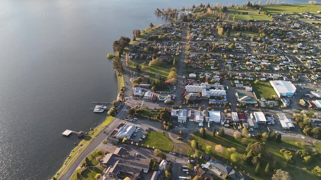Aerial view of Town along Lake Te Anau at sunset, showing lakeside settlement, marina, streets and green spaces at calm sunset evening. Top down shot.