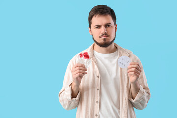 Young man with bloody paper and sad smile on blue background. Hemorrhoids concept