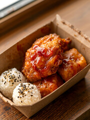 Crispy Fried Chicken Glazed with Spicy Sweet Sauce and Sesame Rice Balls in Cardboard Box. Close-up Gourmet Takeaway Meal on Wooden Table, Delicious Asian Street Food Concept.