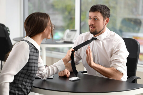 Young woman pulling her male colleague's necktie at desk in office. Harassment concept